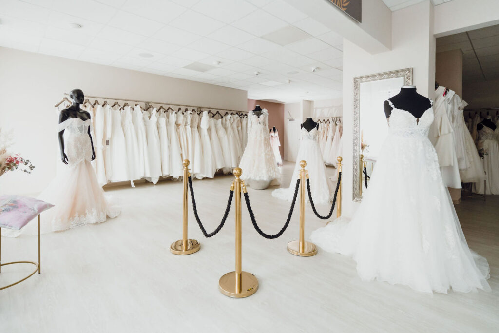 Brautladen-Interieur mit Brautkleidern auf Schaufensterpuppen und Gestellen, großem Spiegel, goldenen Ständern und einer Auswahl an Festmode einschließlich Herrenanzug für den perfekten Look der Hochzeitsgesellschaft.