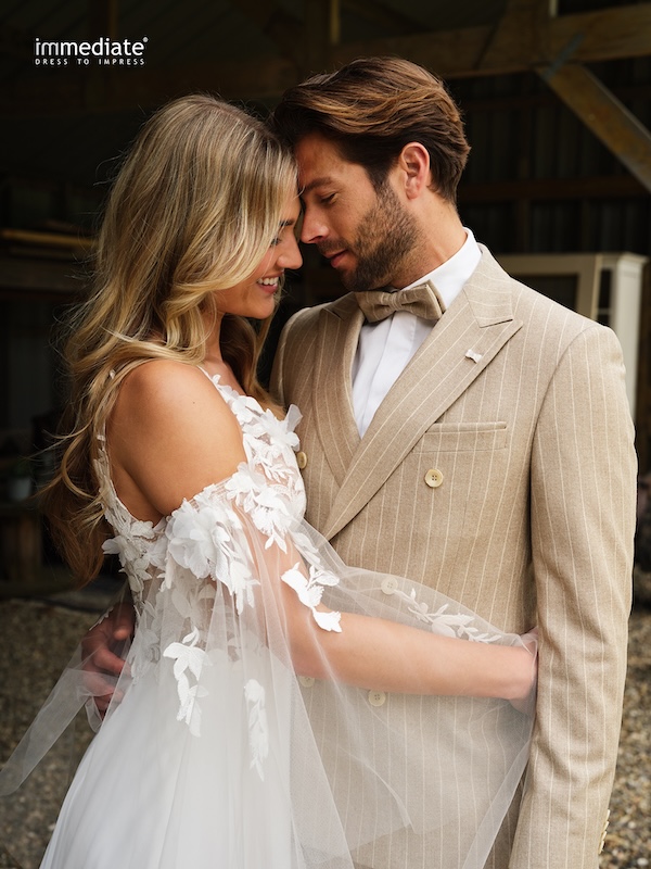 Braut und Bräutigam umarmen sich und lächeln, gekleidet in ein umwerfendes Brautkleid und einen hellbraunen Anzug, und stehen dicht beieinander.
