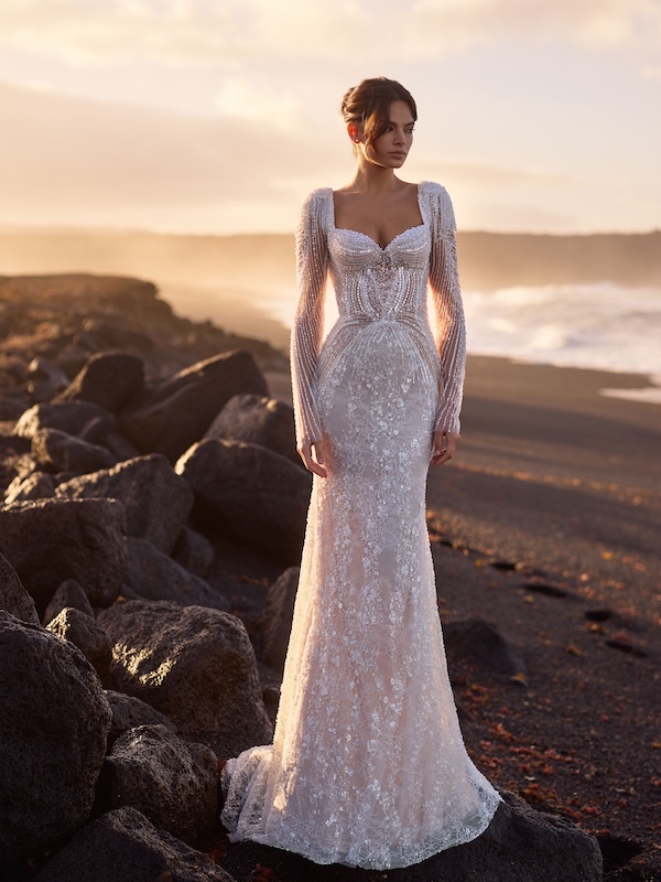 Eine Frau in einem eleganten, langärmeligen Spitzenkleid von Festmode steht bei Sonnenuntergang an einem felsigen Strand.