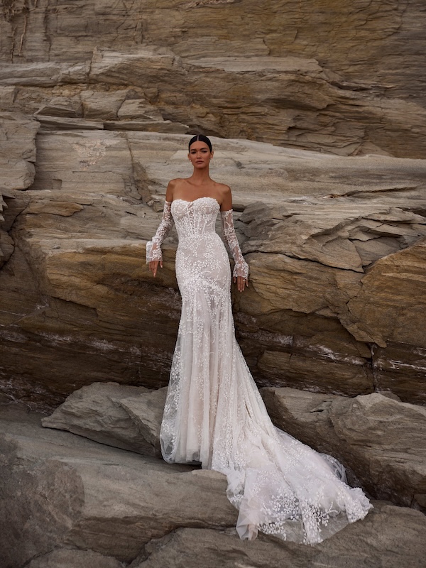 Eine Frau in einem eleganten, schulterfreien Spitzenhochzeitskleid aus einem renommierten Hochzeitshaus steht vor großen Felsen und verkörpert die Essenz der modernen Brautmode.