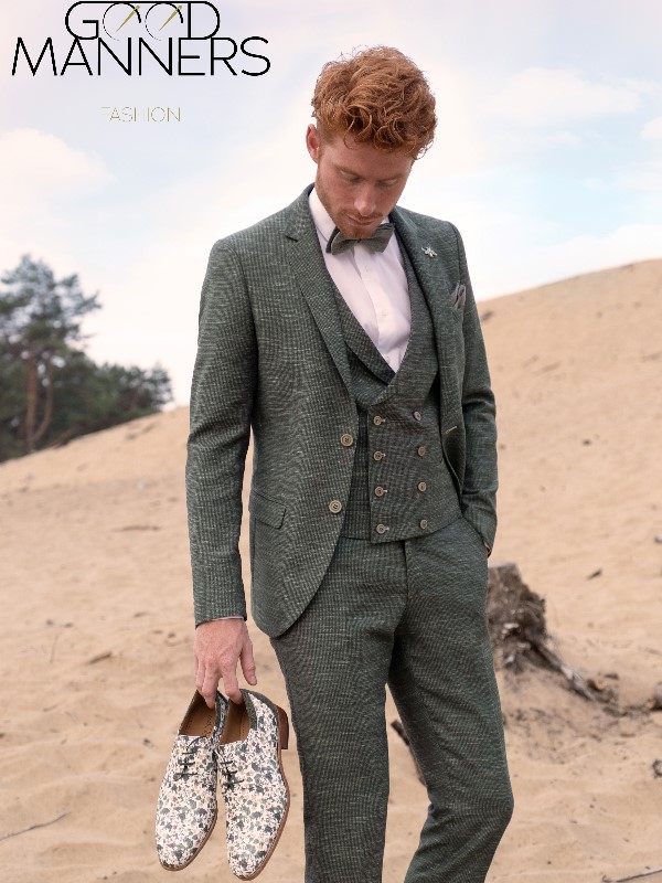 Ein Mann in einem grün karierten Anzug steht auf Sand und hält geblümte Schuhe in der Hand, darüber der Text Good Manners Fashion.