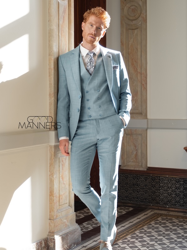 Ein Mann mit rotem Haar in einem hellblauen dreiteiligen Anzug steht in einem sonnendurchfluteten, eleganten Flur mit Marmorverzierungen.