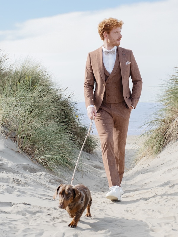 Ein Mann in einem braunen Anzug geht mit seinem Dackel auf einem Sandweg durch grasbewachsene Dünen am Strand spazieren.