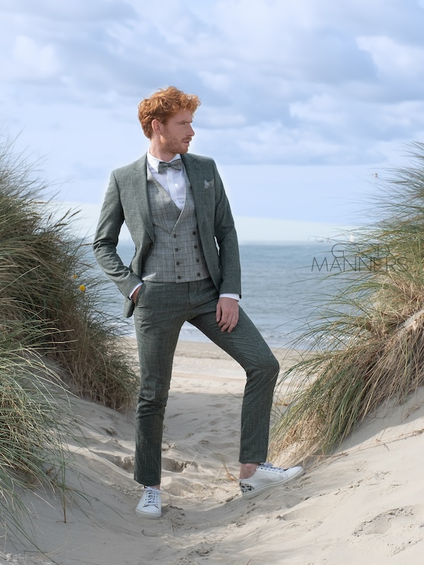 Ein Mann in einem grauen Anzug steht auf einem sandigen Strandweg mit Meer und bewölktem Himmel im Hintergrund.