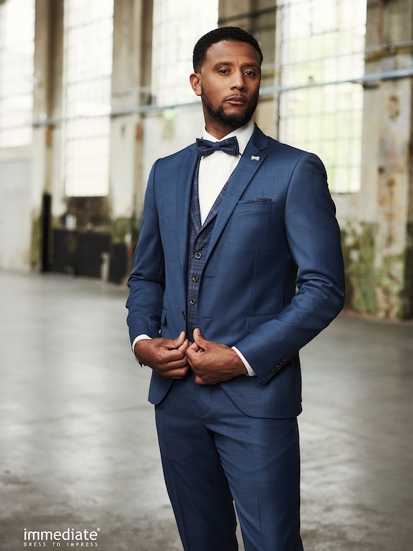 Ein Mann im blauen Anzug und mit Fliege steht in einem geräumigen Raum im Industriestil mit großen Fenstern.