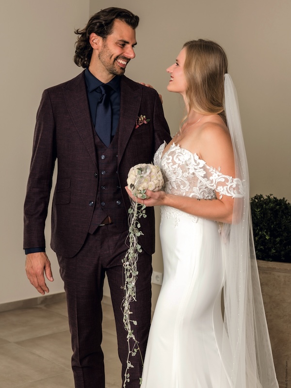 Die Braut in einem weißen Kleid und der Bräutigam in einem dunklen Anzug lächeln sich an und halten einen Blumenstrauß.