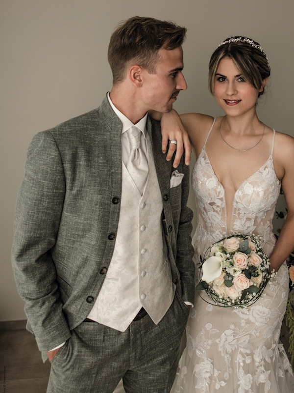 Braut im Spitzenhochzeitskleid mit Blumen in der Hand, stehend neben Bräutigam in grauem Anzug und cremefarbener Weste, lächelnd.