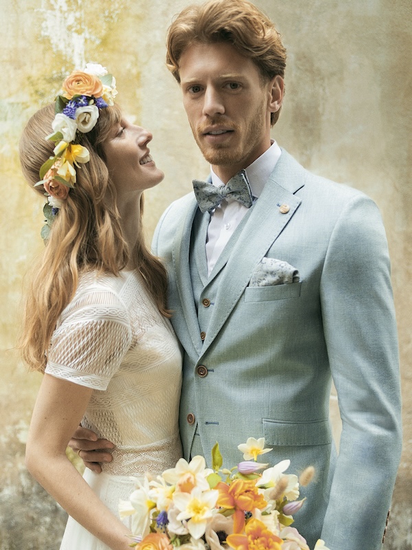 Die Braut im weißen Kleid und mit Blumenkranz lächelt den Bräutigam im hellblauen Anzug an, der einen Blumenstrauß hält.