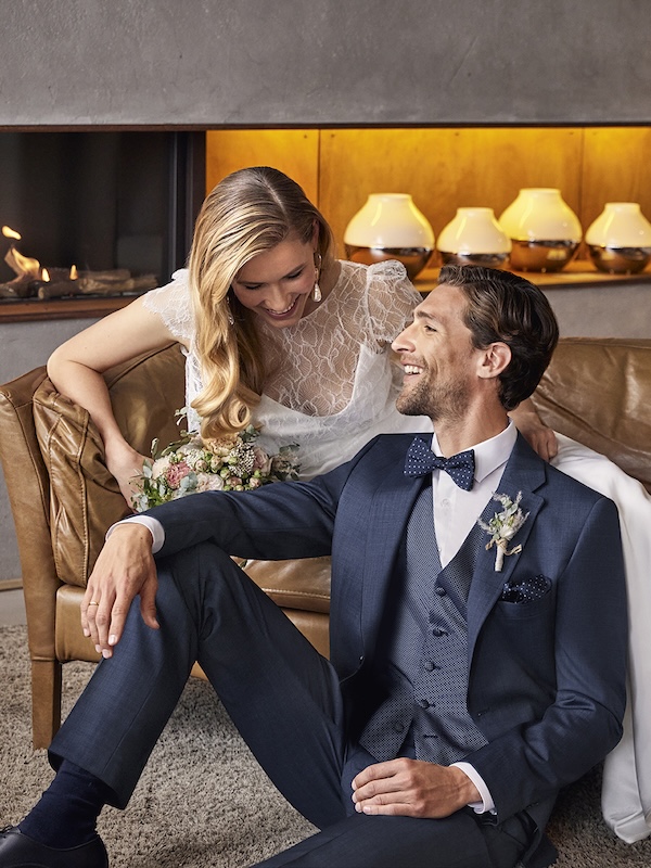 Die Braut im Spitzenkleid und der Bräutigam im blauen Anzug lächeln sich auf einem Ledersofa an und halten einen Blumenstrauß.