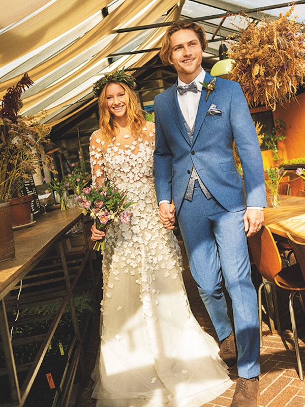 Eine lächelnde Braut in einem geblümten Kleid und ein Bräutigam in einem blauen Anzug halten sich an den Händen und gehen in einem von Pflanzen umgebenen Raum spazieren.