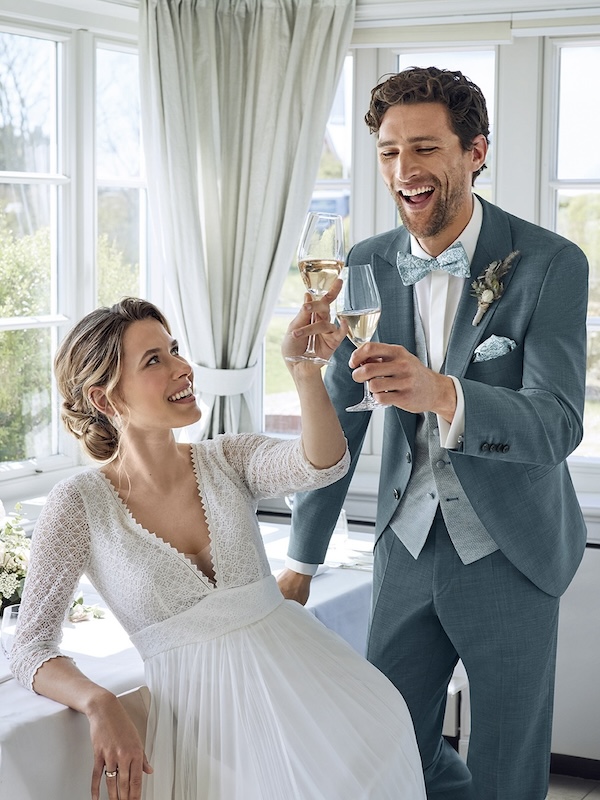 Braut und Bräutigam in Hochzeitskleidung lächeln und stoßen mit Gläsern in einem hellen Raum mit großen Fenstern an.