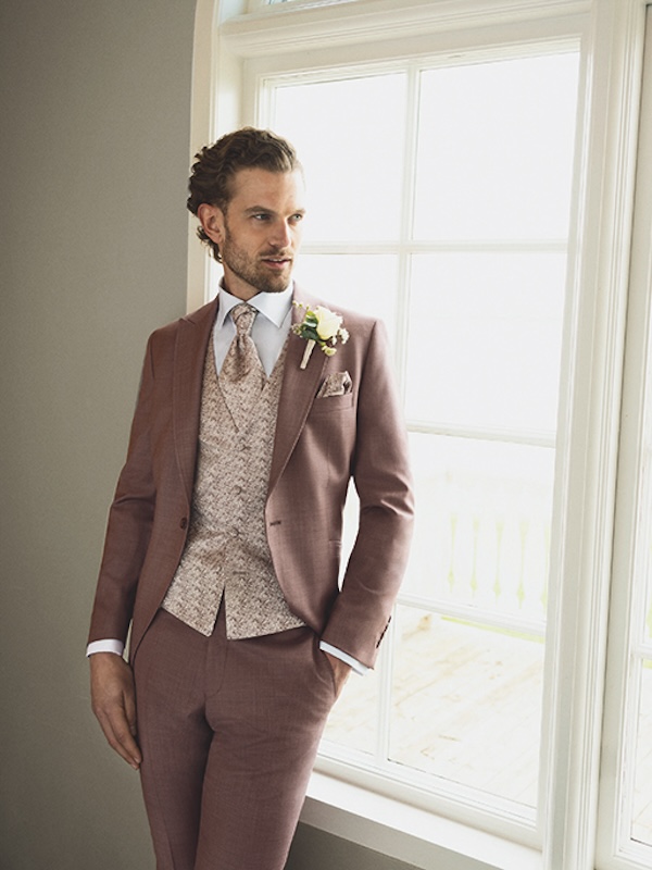 Mann im lilafarbenen Anzug mit gemusterter Weste und Krawatte, der an einem Fenster steht und eine Boutonniere trägt.