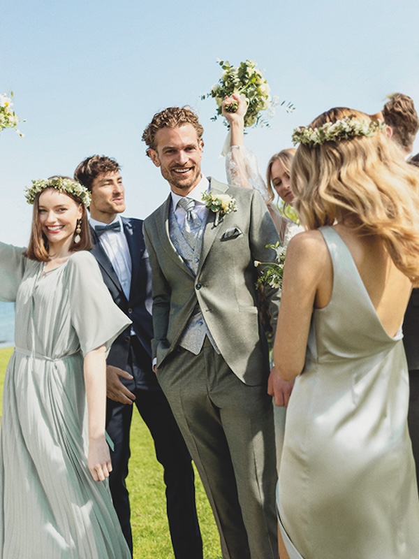 Eine Gruppe formell gekleideter Menschen feiert im Freien eine Hochzeit, lächelnd und mit Blumen in der Hand.